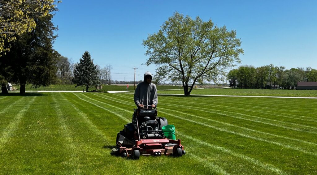 Landcape Lawn Care: lawn care worker riding lawn mower grass mature trees blue skies sunny day